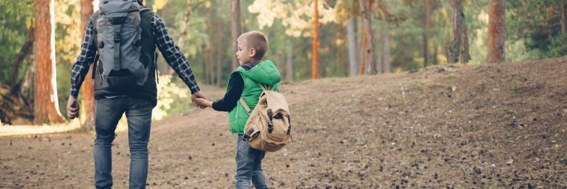 Back view of happy family child and caring father walking in forest with backpacks holding hands and talking, boy is throwing pine cones. Nature, fatherhood and autumn concept.